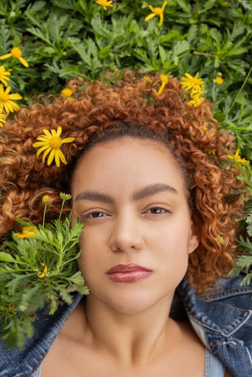 Melissa Oliveira em retrato de beleza, deitada em um jardim de margaridas amarelas, cabelos ruivos cacheados