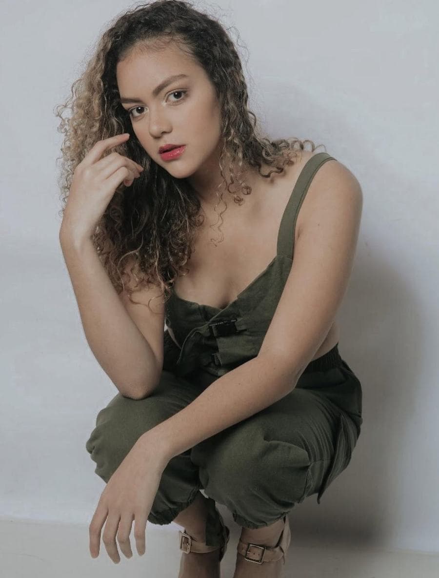 Melissa Oliveira em pose editorial agachada, macacão verde militar, batom vermelho, fundo neutro