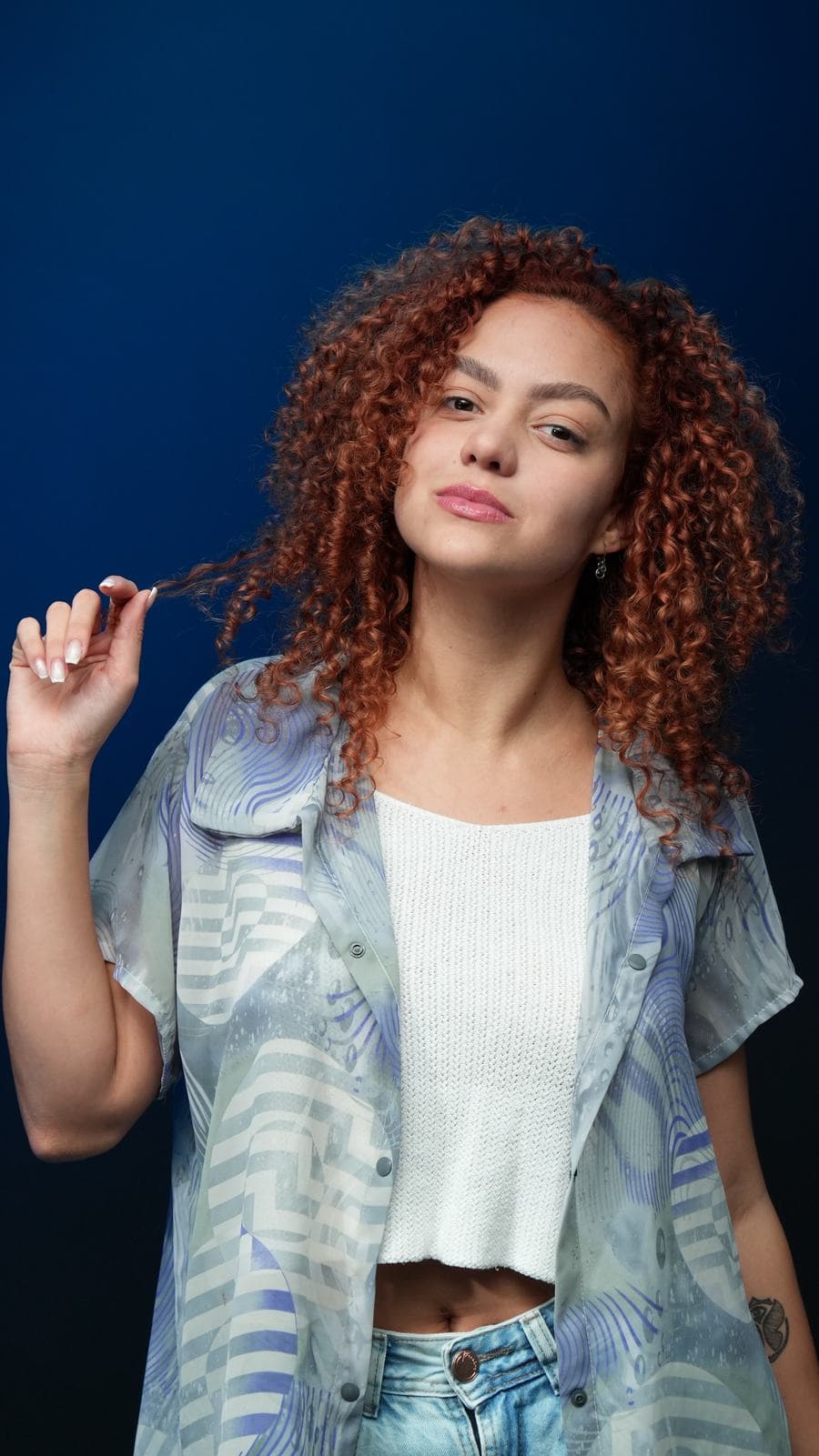 Melissa Oliveira em estúdio com fundo azul, camisa translúcida estampada, brincando com os cachos ruivos