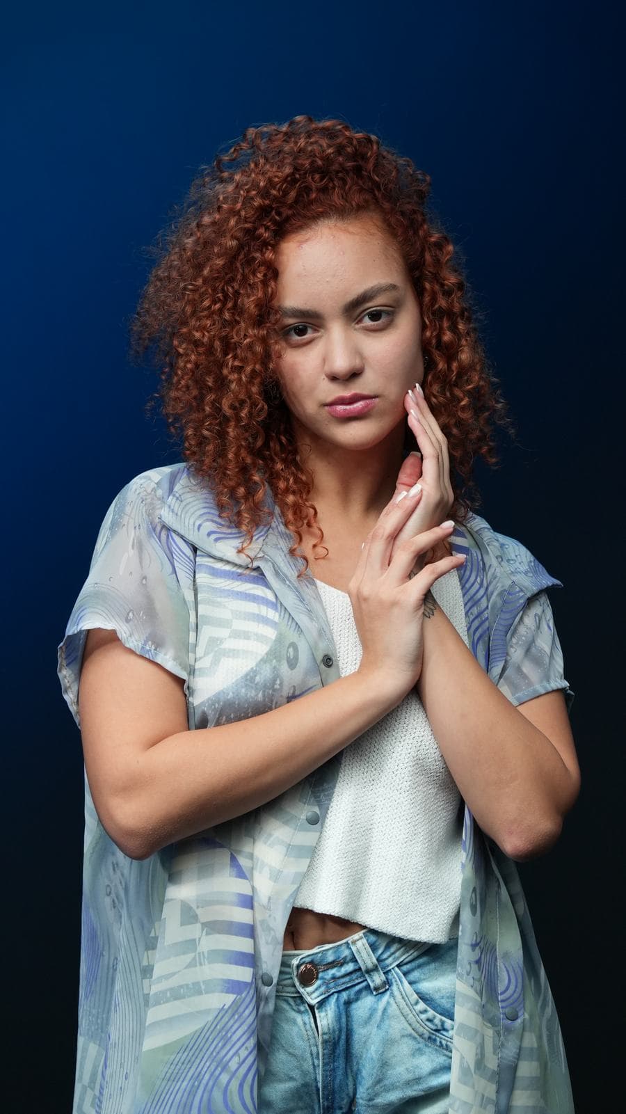 Melissa Oliveira em estúdio com fundo azul, mãos entrelaçadas junto ao rosto, expressão serena
