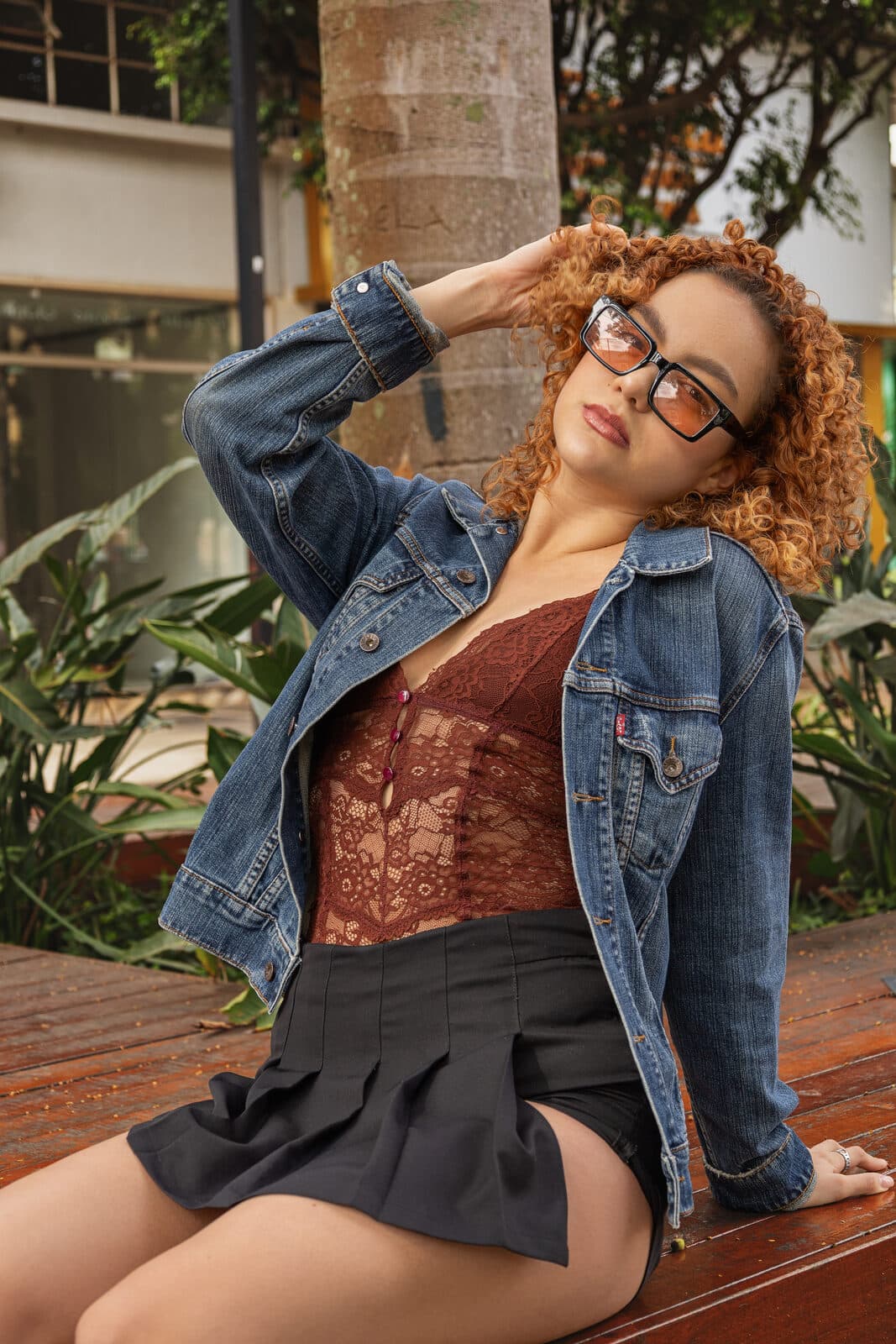 Melissa Oliveira em ensaio lifestyle ao ar livre, jaqueta jeans, top de renda e óculos, sentada em um banco