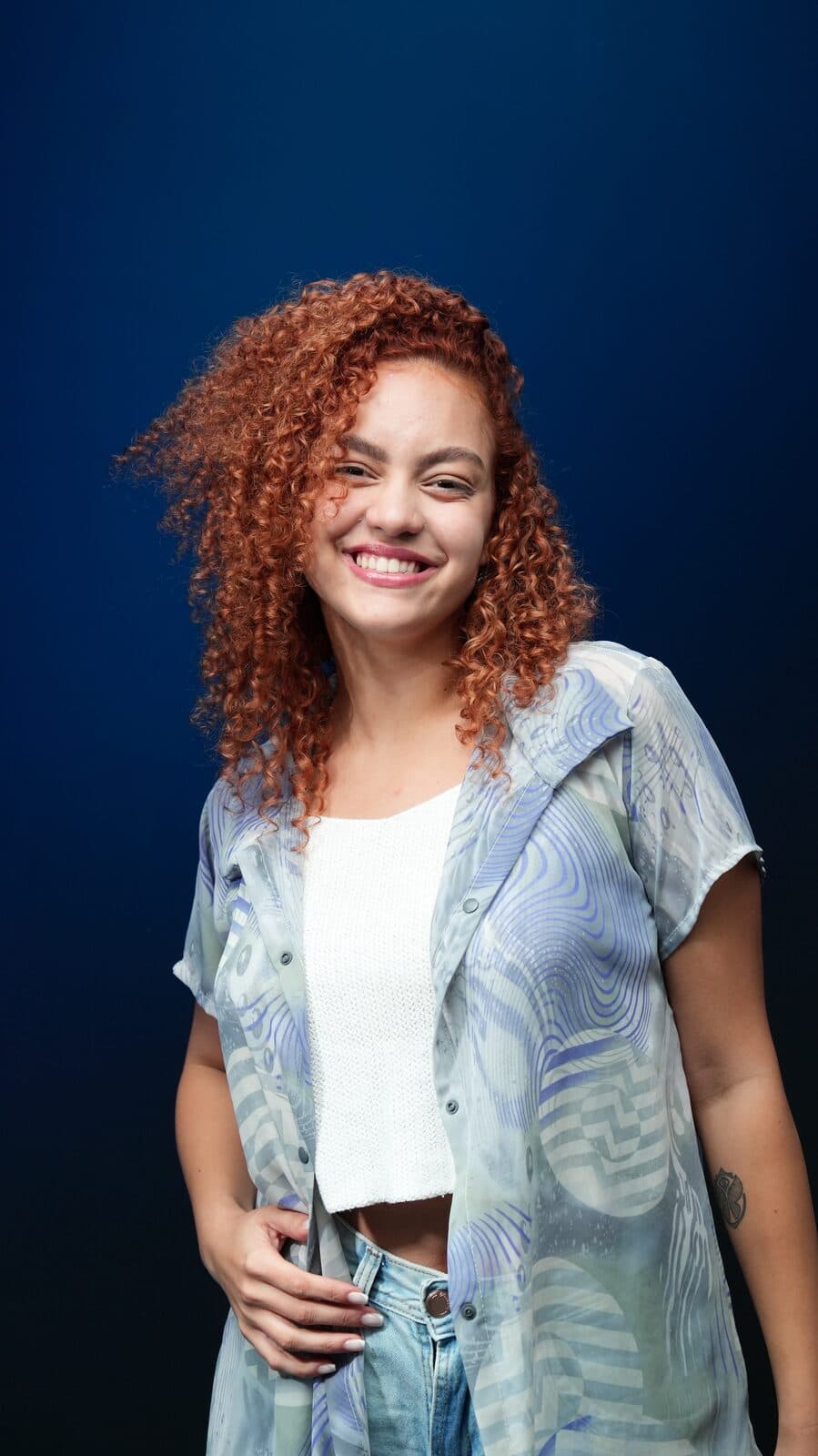 Melissa Oliveira sorrindo em estúdio com fundo azul, camisa translúcida sobre top branco