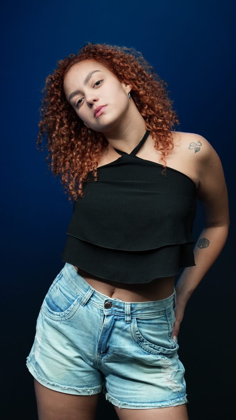 Melissa Oliveira em estúdio com fundo azul, top frente-única preto e shorts jeans, mão na cintura