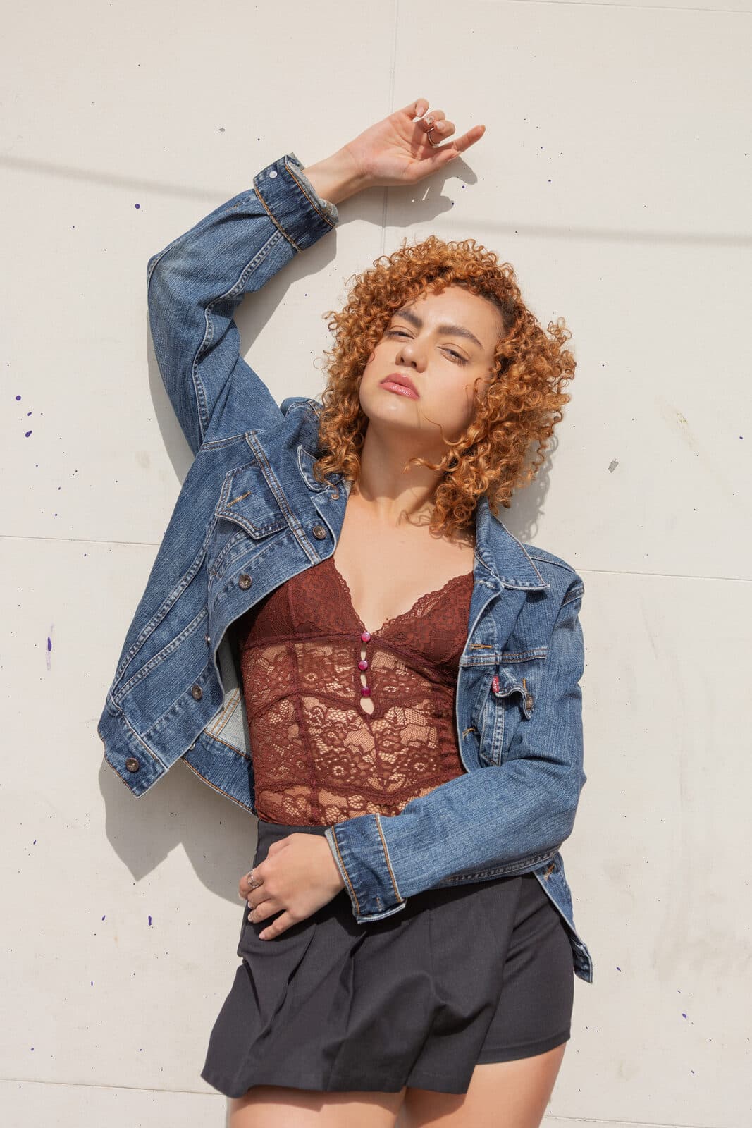 Melissa Oliveira em ensaio ao ar livre sob luz do sol, jaqueta jeans e top de renda, braço erguido junto à parede