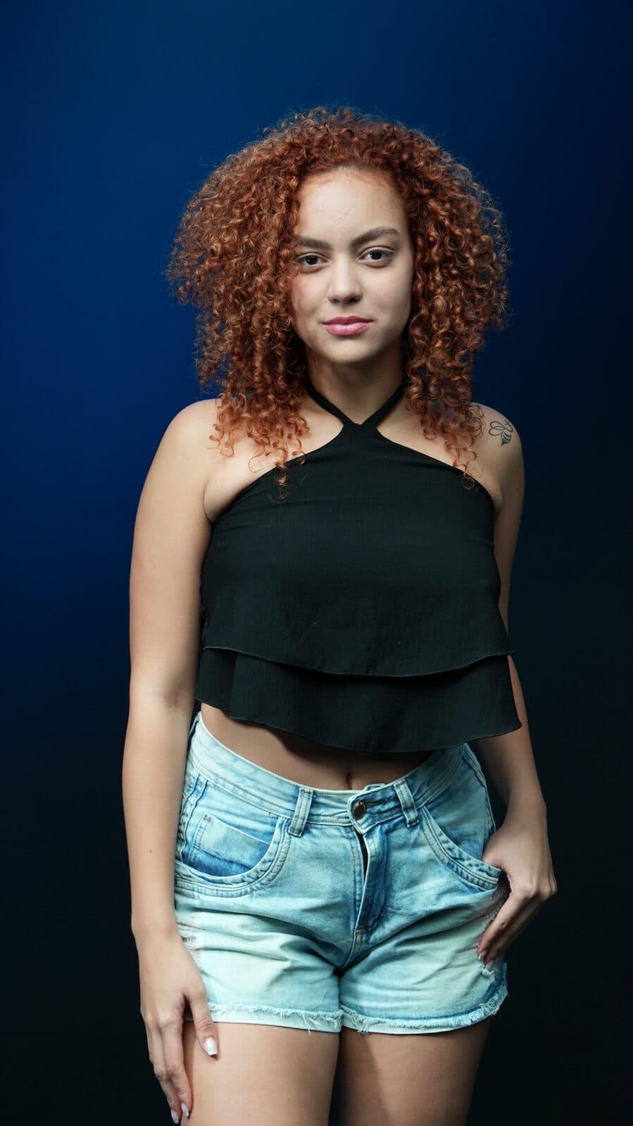 Melissa Oliveira em corpo inteiro em estúdio com fundo azul, top frente-única preto e shorts jeans