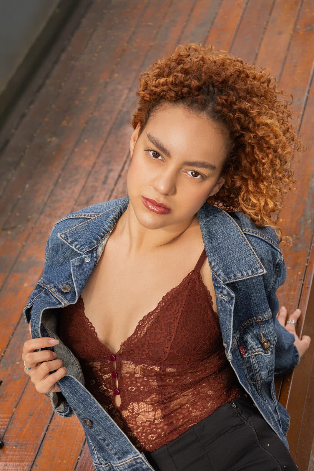 Melissa Oliveira em pose editorial sobre piso de madeira, jaqueta jeans e top de renda, olhando para a câmera