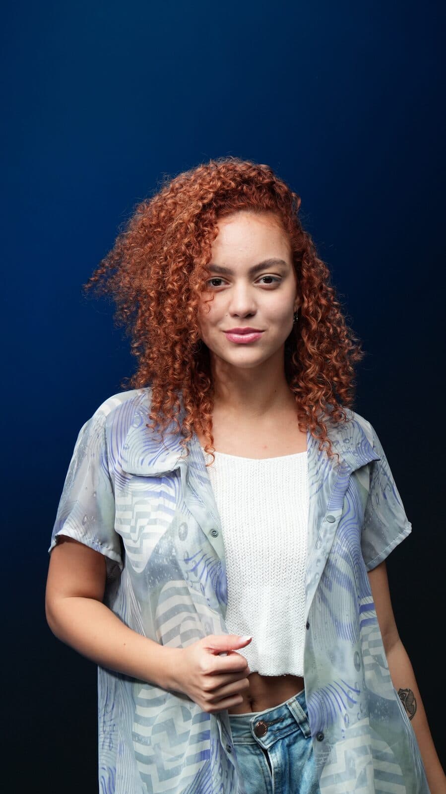 Melissa Oliveira em estúdio com fundo azul, camisa translúcida sobre top branco, expressão serena